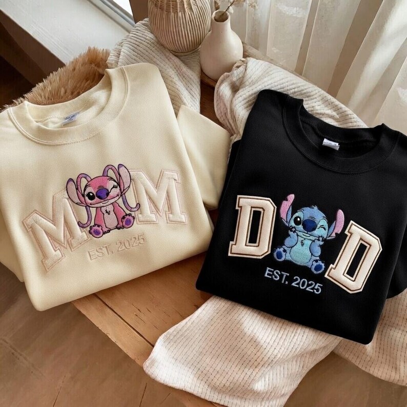 Puede incluir: Dos sudaderas, una crema y otra negra, con dise&ntilde;os de Stitch bordados. La sudadera crema dice "MOM" con un Stitch rosa y "EST. 2025". La sudadera negra dice "DAD" con un Stitch azul y "EST. 2025".