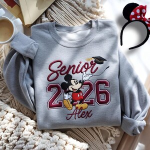 May include: A gray sweatshirt with the text "Senior 2026" and the name "Alex" embroidered in burgundy. A cartoon Mickey Mouse character is featured, wearing a graduation cap. A black headband with red polka dots is also visible.