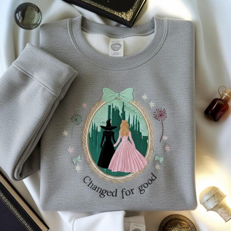 May include: A gray sweatshirt with an embroidered design featuring two figures in front of a green cityscape, with the text "Changed for good". The design is framed by a light green bow and a decorative border. Additional embroidered elements include stars and bows.