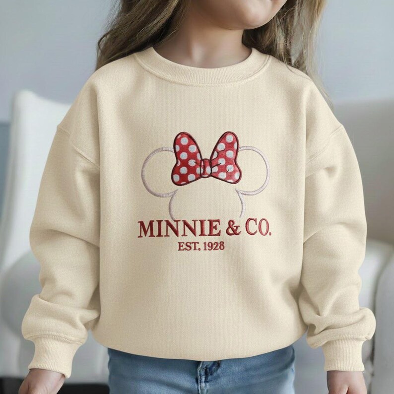 Op de afbeelding: Cr&egrave;mekleurige sweatshirt met een rode polka-dot strik en Minnie Mouse oren. De tekst "MINNIE & CO. EST. 1928" is rood geborduurd. Een comfortabele top met lange mouwen voor kinderen.