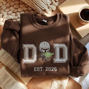 May include: A brown sweatshirt with the word "DAD" in white block letters, featuring an embroidered image of a Mandalorian character and Grogu. The text "EST. 2026" is below the graphic. The sweatshirt is folded on a neutral surface.