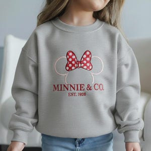 Op de afbeelding: Een grijze sweatshirt met een rode strik met witte stippen en de tekst "MINNIE & CO. EST. 1928" in het rood. Het ontwerp bevat een roze omtrek van Minnie Mouses hoofd. Kinderkleding.