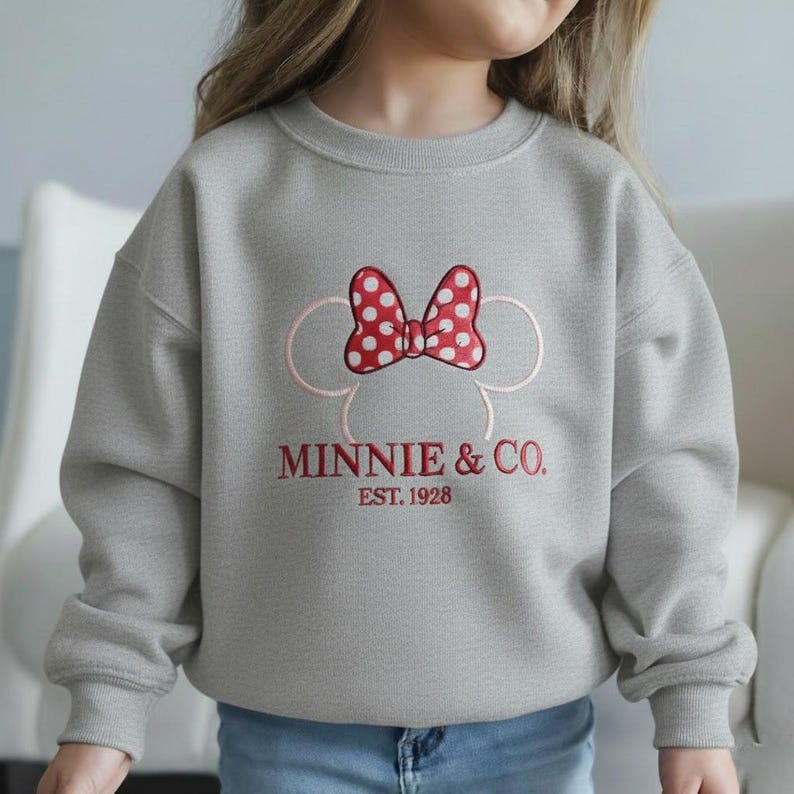 Op de afbeelding: Een grijze sweatshirt met een rode strik met witte stippen en de tekst "MINNIE & CO. EST. 1928" in het rood. Het ontwerp bevat een roze omtrek van Minnie Mouses hoofd. Kinderkleding.