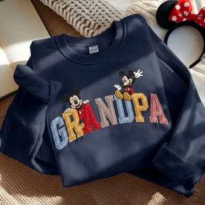 May include: A navy blue sweatshirt featuring the word "GRANDPA" embroidered in colourful letters, with two Mickey Mouse figures. A Minnie Mouse ear headband with a red polka dot bow is visible in the background.