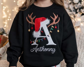May include: Black sweatshirt with an embroidered Christmas design. The design features a large white letter "A" with a red Santa hat, reindeer antlers, and string lights. The name "Anthonny" is embroidered below the letter in a cursive font. The sweatshirt is part of a holiday collection.
