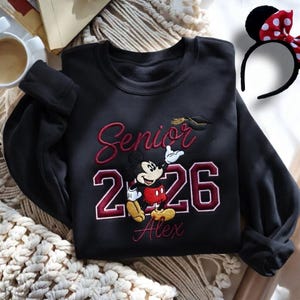 May include: Black sweatshirt with embroidered "Senior 2026" text, Mickey Mouse graphic, and the name "Alex". A graduation cap is embroidered above the numbers. A black headband with Minnie Mouse ears and a red polka dot bow is also visible.