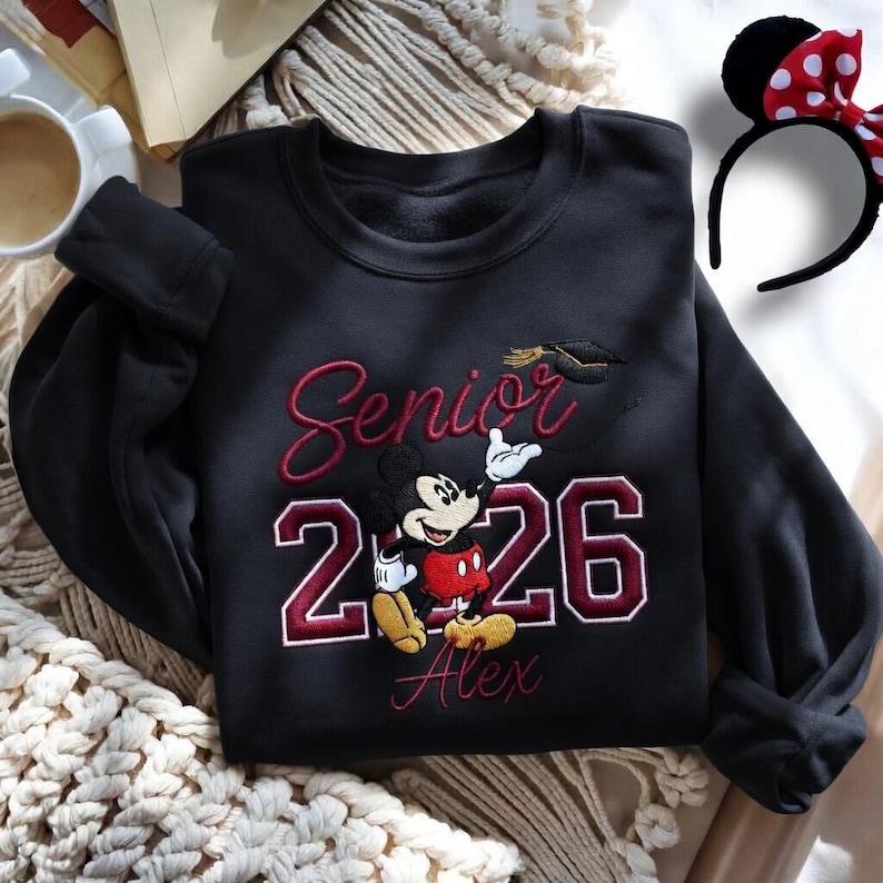 May include: Black sweatshirt with embroidered "Senior 2026" text, Mickey Mouse graphic, and the name "Alex". A graduation cap is embroidered above the numbers. A black headband with Minnie Mouse ears and a red polka dot bow is also visible.
