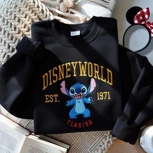 May include: Black sweatshirt with "DISNEYWORLD" in gold lettering, featuring a blue Stitch character and the text "EST. 1971 FLORIDA". A Minnie Mouse ear headband is visible in the background.