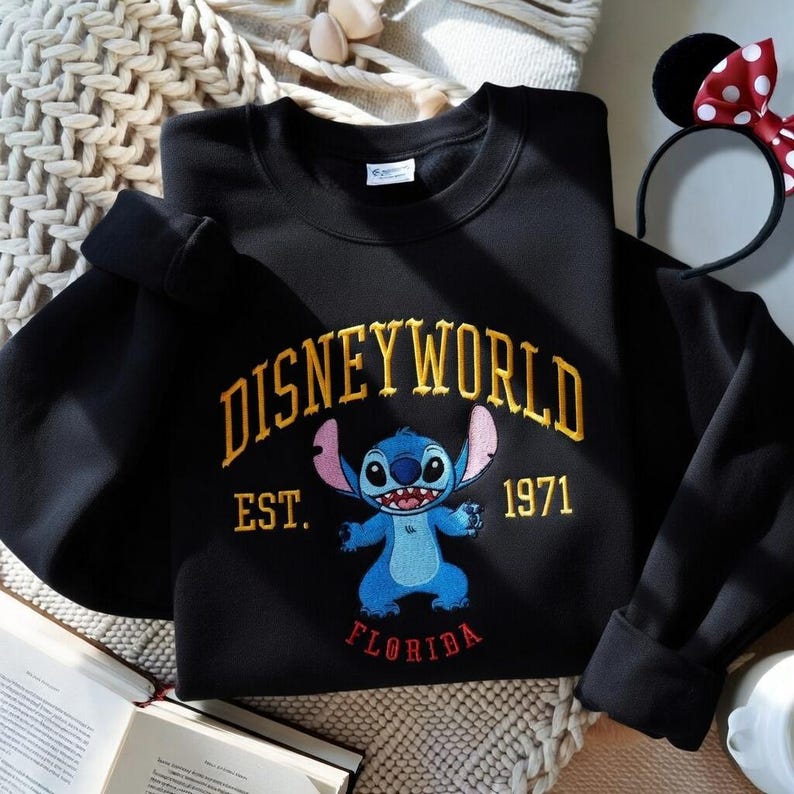 May include: Black sweatshirt with "DISNEYWORLD" in gold lettering, featuring a blue Stitch character and the text "EST. 1971 FLORIDA". A Minnie Mouse ear headband is visible in the background.