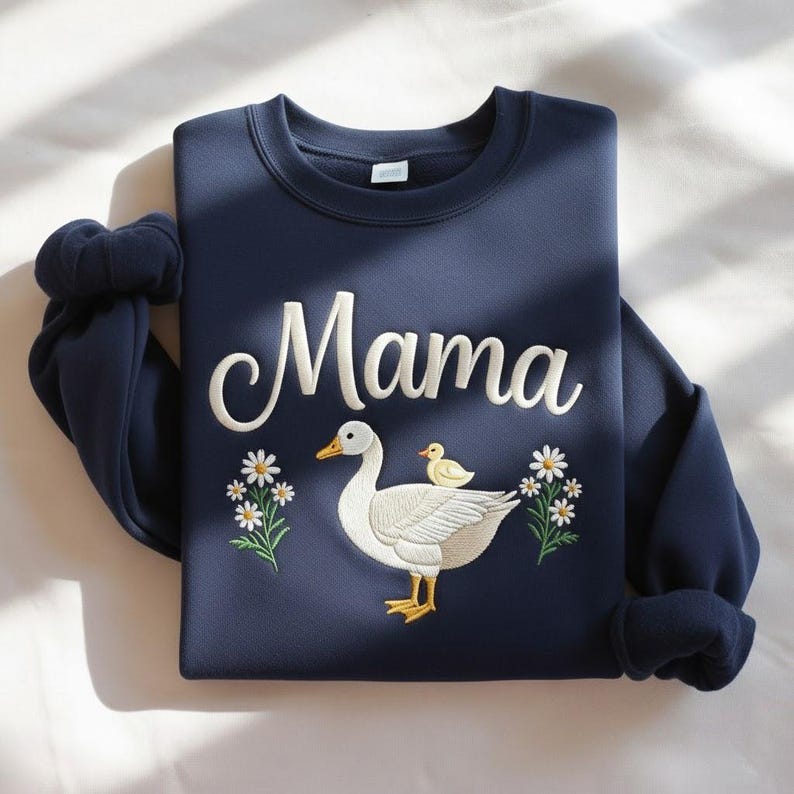 May include: Navy blue sweatshirt with the word "Mama" in white script. The design features a white goose with a yellow duckling, surrounded by white daisies with green stems and leaves. The sweatshirt has a crew neck and long sleeves.