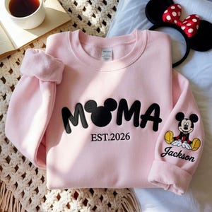 May include: A light pink sweatshirt with the word "MOMA" in black, featuring a Mickey Mouse head in place of the "O". The text "EST.2026" is below the word "MOMA". The sleeve has a Mickey Mouse graphic and the name "Jackson".