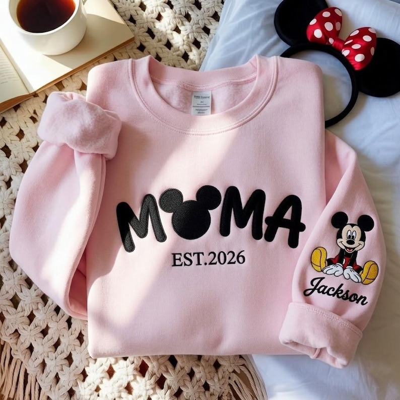 May include: A light pink sweatshirt with the word "MOMA" in black, featuring a Mickey Mouse head in place of the "O". The text "EST.2026" is below the word "MOMA". The sleeve has a Mickey Mouse graphic and the name "Jackson".