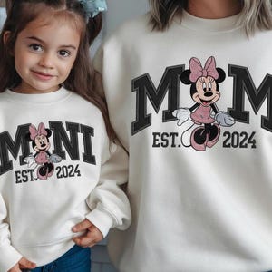 May include: Off-white sweatshirts with matching designs. One sweatshirt says "MOM EST. 2024" with a Minnie Mouse graphic. The other says "MINI EST. 2024" with a similar Minnie Mouse graphic. Both sweatshirts have black block lettering.