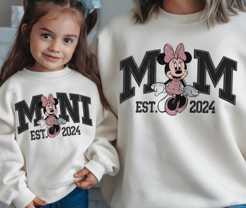 May include: Off-white sweatshirts with matching designs. One sweatshirt says "MOM EST. 2024" with a Minnie Mouse graphic. The other says "MINI EST. 2024" with a similar Minnie Mouse graphic. Both sweatshirts have black block lettering.