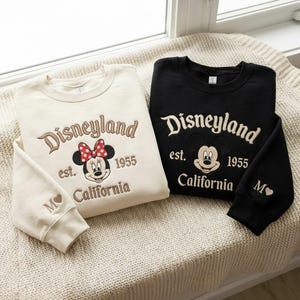 Puede incluir: Dos sudaderas de cuello redondo, una crema y otra negra, bordadas con "Disneyland est. 1955 California" y un gr&aacute;fico de Minnie Mouse o Mickey Mouse. La sudadera crema tiene un lazo rojo y la letra "M" con un coraz&oacute;n en la manga.
