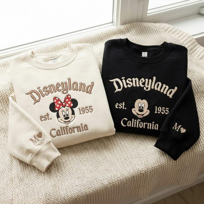 Puede incluir: Dos sudaderas de cuello redondo, una crema y otra negra, bordadas con "Disneyland est. 1955 California" y un gr&aacute;fico de Minnie Mouse o Mickey Mouse. La sudadera crema tiene un lazo rojo y la letra "M" con un coraz&oacute;n en la manga.