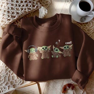 May include: A brown sweatshirt featuring a graphic of four cartoon characters. The characters are green with large ears and brown robes. One character wears headphones, and another has a question mark above its head. The word "cool!" is printed on the sweatshirt.