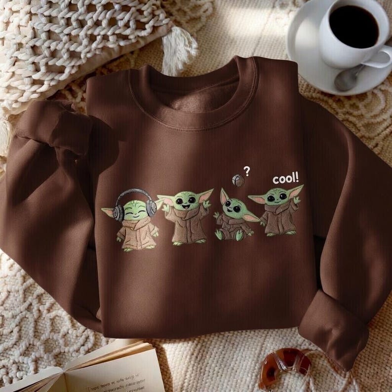 May include: A brown sweatshirt featuring a graphic of four cartoon characters. The characters are green with large ears and brown robes. One character wears headphones, and another has a question mark above its head. The word "cool!" is printed on the sweatshirt.