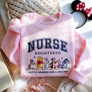 May include: A light pink sweatshirt with the word "NURSE" in navy blue collegiate lettering. Below, the word "REGISTERED" is displayed. The bottom of the sweatshirt features cartoon characters and the phrase "A LITTLE KINDNESS GOES A LONG WAY."