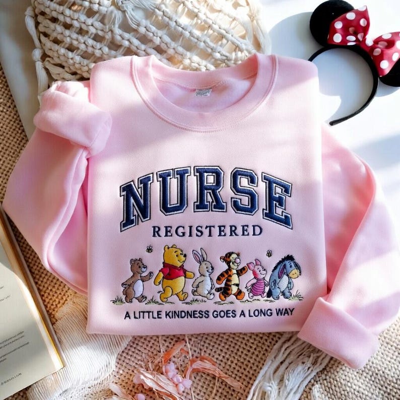 May include: A light pink sweatshirt with the word "NURSE" in navy blue collegiate lettering. Below, the word "REGISTERED" is displayed. The bottom of the sweatshirt features cartoon characters and the phrase "A LITTLE KINDNESS GOES A LONG WAY."