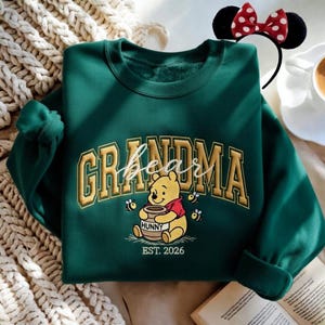 May include: A dark green sweatshirt with the word "GRANDMA" in gold lettering and a Winnie the Pooh graphic. The graphic includes the word "bear" in script, Pooh holding a honey pot, bees, and the text "EST. 2026". A Minnie Mouse ear headband is in the background.