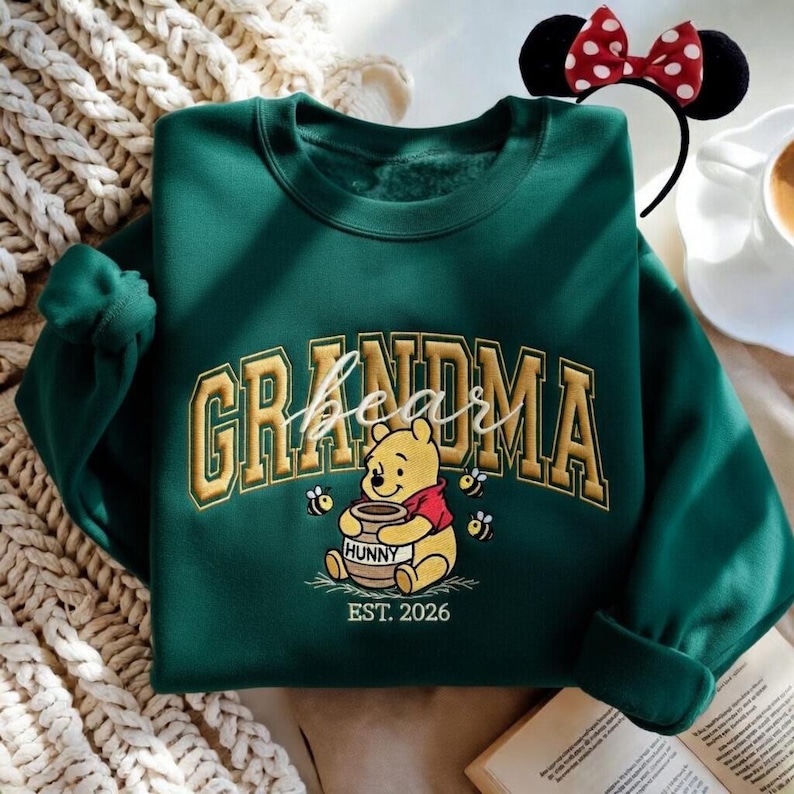 May include: A dark green sweatshirt with the word "GRANDMA" in gold lettering and a Winnie the Pooh graphic. The graphic includes the word "bear" in script, Pooh holding a honey pot, bees, and the text "EST. 2026". A Minnie Mouse ear headband is in the background.