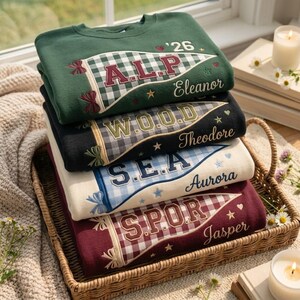 Könnte beinhalten: Ein Stapel aus vier Rundhals-Sweatshirts in Dunkelgrün, Schwarz, Creme und Kastanienbraun. Jedes zeigt einen gestickten Wimpel mit Vichy-Muster, einen Namen sowie kleine Sterne und Herzen. Die Namen sind Eleanor, Theodore, Aurora und Jasper.