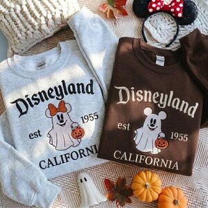 May include: A light gray sweatshirt and a dark brown sweatshirt, both featuring the text Disneyland est 1955 CALIFORNIA and a graphic of a ghost character holding a pumpkin. The gray sweatshirt ghost wears an orange bow, while the brown one does not.