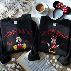 May include: Two black sweatshirts with the words "DISNEY WORLD EST 1971" embroidered in red. One features Mickey Mouse, the other Minnie Mouse. A Minnie Mouse ear headband and a cup of coffee are also visible.
