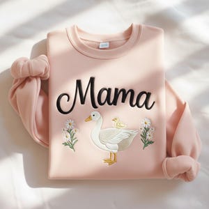 May include: A light pink sweatshirt with the word "Mama" embroidered in black script. Below the text is an embroidered design of a white goose with a yellow beak and feet, and a yellow chick. White daisies are embroidered on either side.