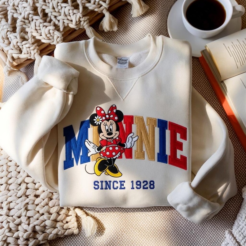 K&ouml;nnte beinhalten: Cremefarbenes Sweatshirt mit Minnie Mouse-Grafik. Das Design zeigt das Wort "MINNIE" in Rot, Blau und Gold, mit Minnie Mouse in einem roten gepunkteten Kleid. Der Text "SINCE 1928" steht darunter.