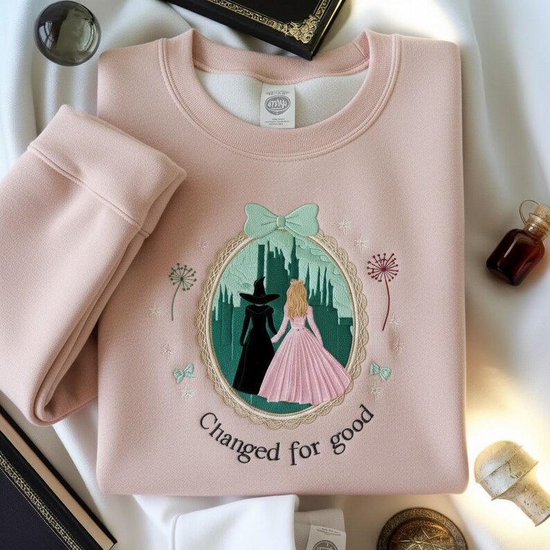 May include: A light pink sweatshirt with an embroidered design featuring two figures in front of a green cityscape. The design is framed by a light green bow and the words "Changed for good".