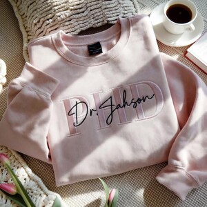 May include: A light pink sweatshirt with the embroidered text "PHD Dr. Jahson" in a matching pink color and black script. The sweatshirt has a crew neck and long sleeves. A cup of coffee and a book are in the background.