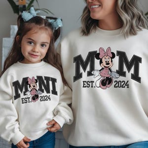 May include: Matching cream-colored sweatshirts featuring Minnie Mouse. One sweatshirt says "MOM EST. 2024" in black block letters with a Minnie Mouse graphic. The other says "MINI EST. 2024" with a similar design. Both sweatshirts are worn by a mother and child.