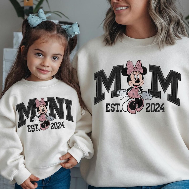 May include: Matching cream-colored sweatshirts featuring Minnie Mouse. One sweatshirt says "MOM EST. 2024" in black block letters with a Minnie Mouse graphic. The other says "MINI EST. 2024" with a similar design. Both sweatshirts are worn by a mother and child.