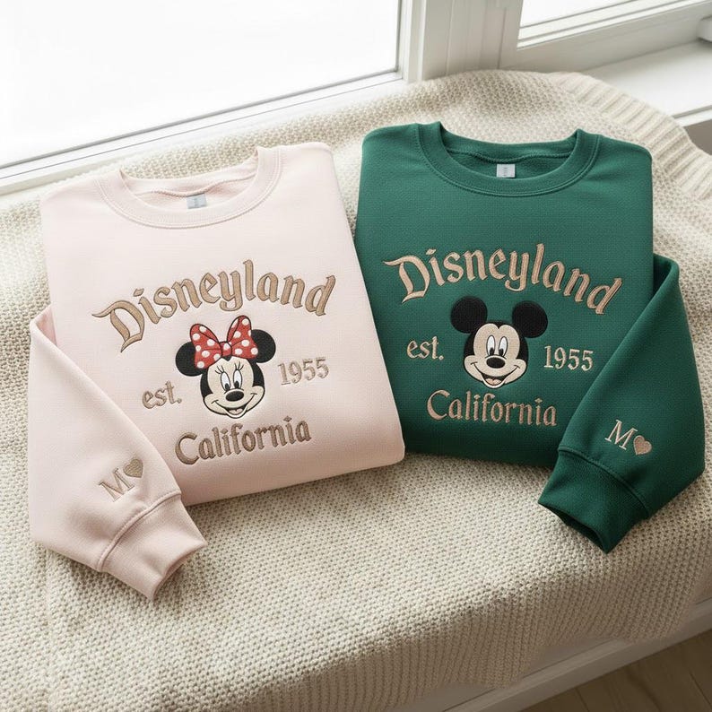 Puede incluir: Dos sudaderas, una rosa claro y otra verde, con el logotipo de Disneyland y Mickey y Minnie Mouse. Las sudaderas tienen el texto "Disneyland est. 1955 California". La sudadera rosa claro tiene un dise&ntilde;o de Minnie Mouse, y la verde tiene un dise&ntilde;o de Mickey Mouse.
