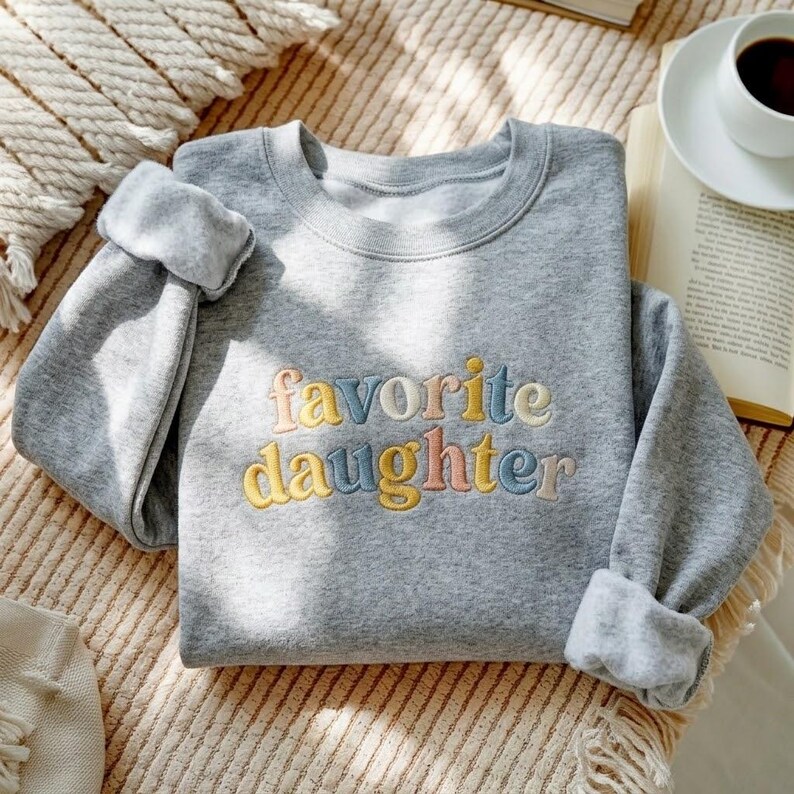 May include: A grey sweatshirt featuring the embroidered words "favorite daughter" in pastel shades. The sweatshirt has a crew neck and rolled-up sleeves. The image is taken from an overhead perspective.