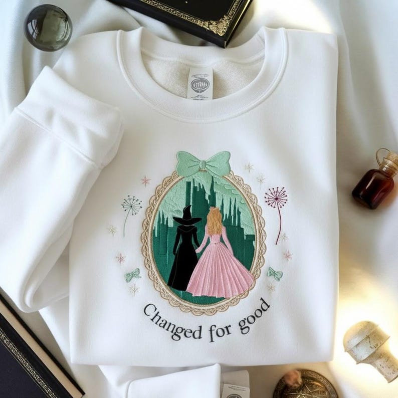 May include: White sweatshirt with an embroidered design featuring two figures in front of a green cityscape, with the text "Changed for good". The design is framed by a scalloped border and a mint green bow.