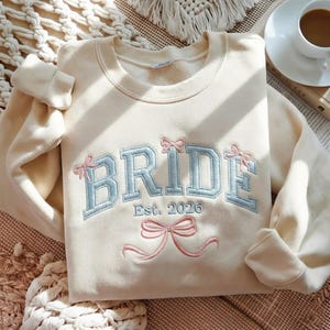 May include: Cream-colored sweatshirt with the word "BRIDE" embroidered in light blue, accented with pink bows. The text "Est. 2026" is below the main word. The sweatshirt is folded on a textured surface, suggesting a bridal theme.