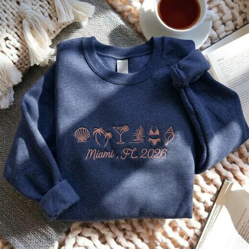 Peut inclure: Un sweat-shirt bleu marine &agrave; col rond. Le devant pr&eacute;sente un motif corail de coquillages, de palmiers, d'un cocktail et d'un bikini, avec le texte "Miami, FL 2026". Le sweat-shirt est pli&eacute; sur une surface textur&eacute;e.