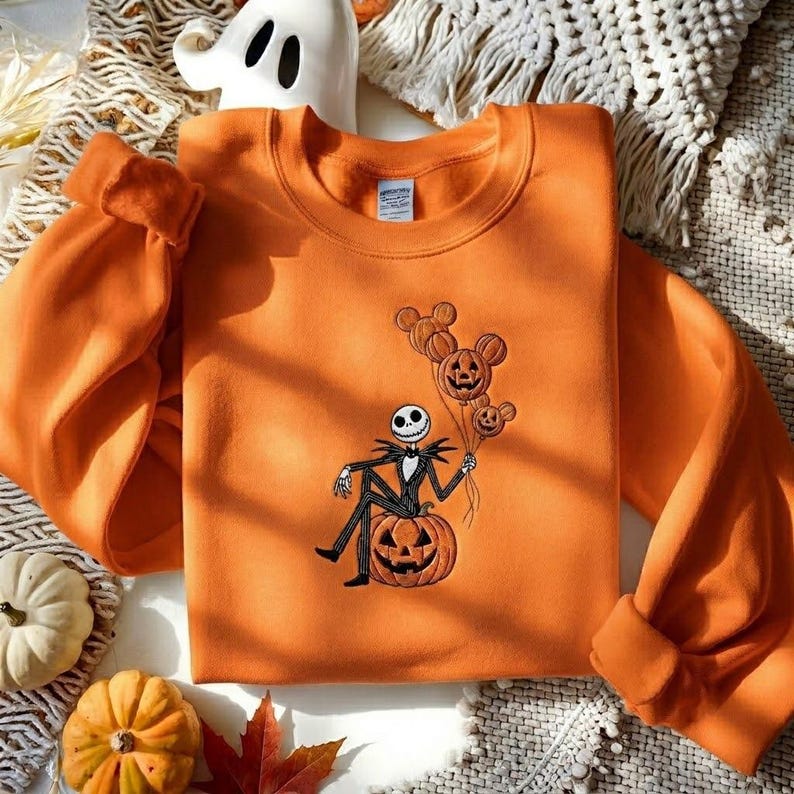 May include: An orange crewneck sweatshirt featuring an embroidered design of a skeleton character sitting on a large jack-o'-lantern while holding three pumpkin-shaped balloons. The embroidery uses black, white, and orange thread colors.