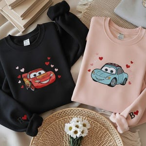 Op de afbeelding: Twee sweatshirts, één zwart en één roze, elk geborduurd met een cartoon auto-ontwerp en kleine rode hartjes. Het zwarte sweatshirt heeft een rode auto, terwijl de roze een blauwe auto heeft. De mouwen hebben geborduurde initialen.