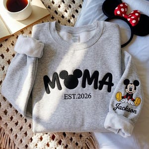 May include: A light gray sweatshirt with the word "MOMA" in black, featuring a Mickey Mouse head as the "O". The text "EST.2026" is below the word. The sleeve has a Mickey Mouse graphic and the name "Jackson".