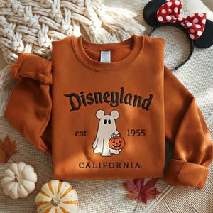 Pode incluir: Uma camisola de manga comprida em cor de laranja queimado com texto bordado a preto que diz Disneyland California. O gr&aacute;fico central mostra um fantasma branco com orelhas de rato a segurar uma ab&oacute;bora laranja, com o texto est 1955.