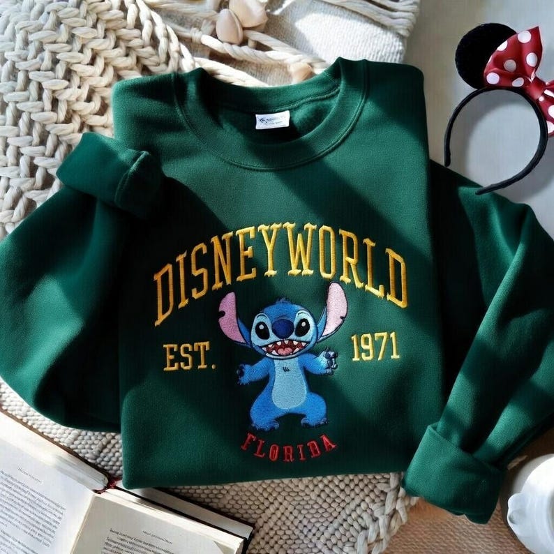 May include: A dark green sweatshirt featuring the word "DISNEYWORLD" in gold lettering, with "EST. 1971" below. A blue cartoon character is in the center, with "FLORIDA" below. A Minnie Mouse ear headband is visible.