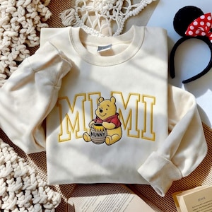 以下が含まれることがあります： クリーム色のスウェットシャツ。黄色いブロック体で「MIMI」の文字が刺繍されています。文字の下には、蜂蜜の壺を持った漫画のくまのプーさんのキャラクターが中央に配置されています。赤と白の水玉模様のリボンが付いた黒いヘアバンドが背景にあります。