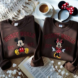 May include: Two brown Disney World sweatshirts with embroidered Mickey and Minnie Mouse designs. The sweatshirts have the text "Disney World EST 1971" in red. A Minnie Mouse ear headband and a cup of coffee are also in the image.