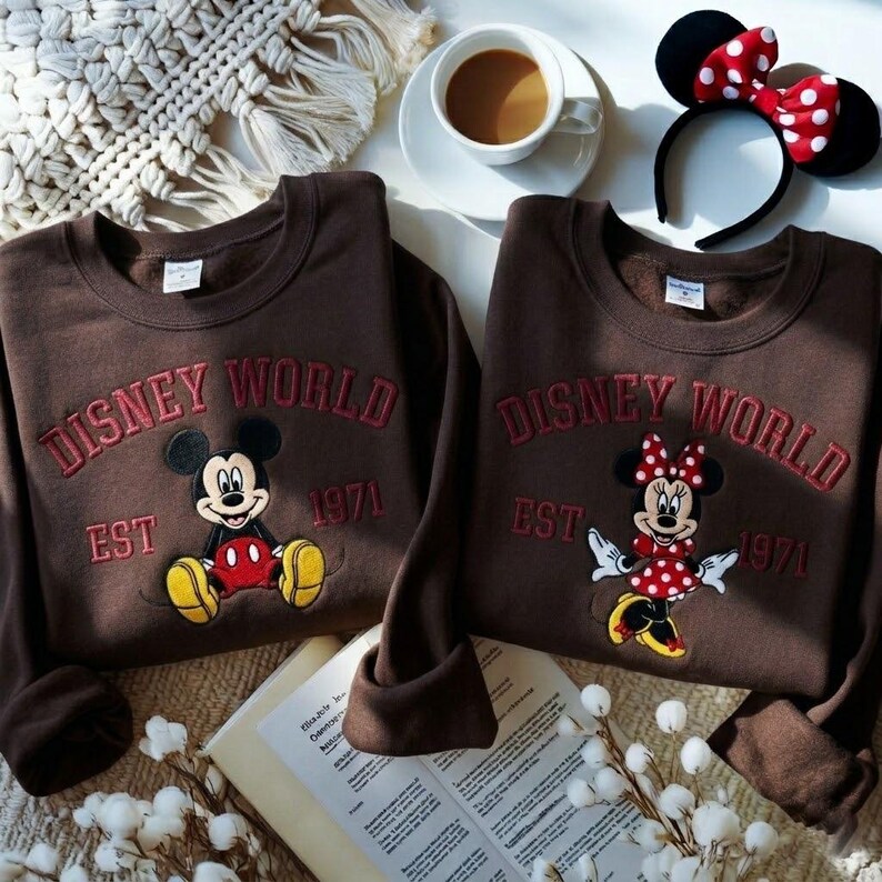 May include: Two brown Disney World sweatshirts with embroidered Mickey and Minnie Mouse designs. The sweatshirts have the text "Disney World EST 1971" in red. A Minnie Mouse ear headband and a cup of coffee are also in the image.