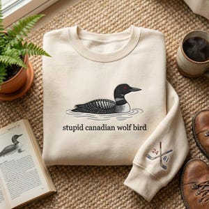 May include: Cream-colored sweatshirt featuring a black and white illustration of a loon on water, with the text "stupid canadian wolf bird" below. The sleeve has embroidered hockey sticks and numbers 24 and 81.