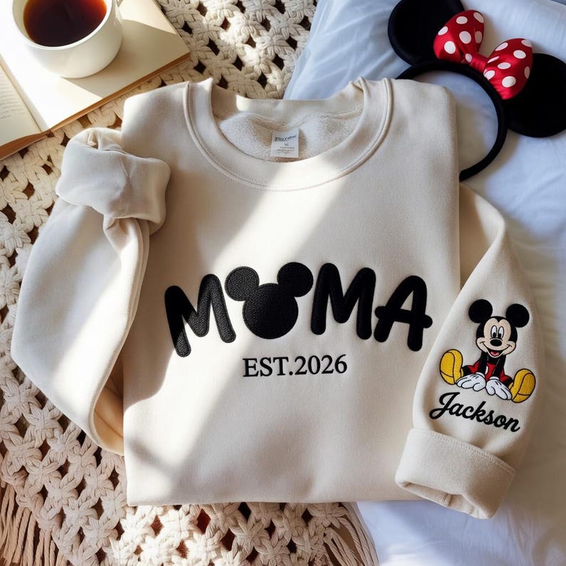 May include: Cream-colored sweatshirt with the word "MAMA" in black, featuring a Mickey Mouse head in place of the "O". The text "EST.2026" is below. The right sleeve has a Mickey Mouse graphic and the name "Jackson".